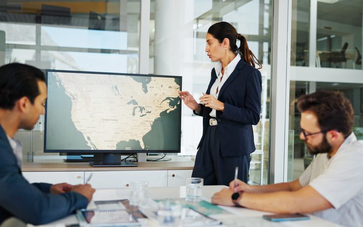 A presenter in a navy suit explains a world map on a monitor to two colleagues in a glass-walled office conference room.