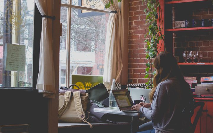 A person with long hair working on a laptop at a small table in a cozy, brick-walled cafe with large window, curtains, and indoor plants.