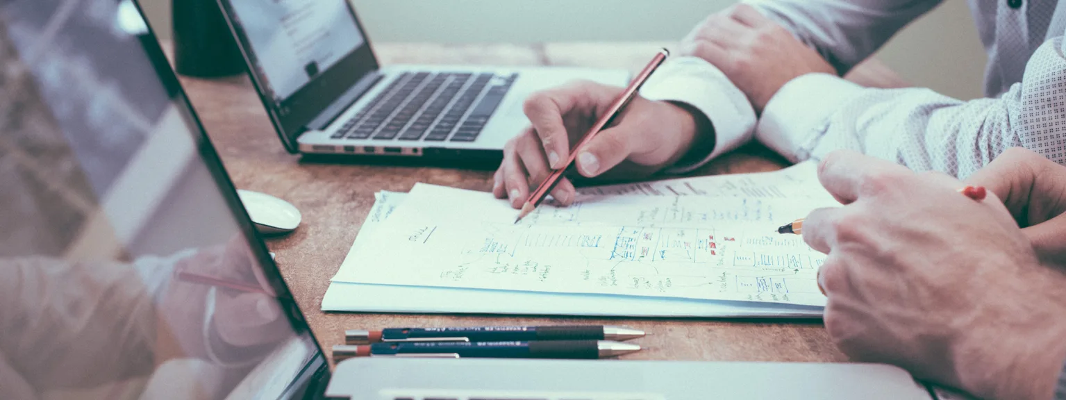 Due persone discutono di un progetto con appunti e due laptop su un tavolo di legno.