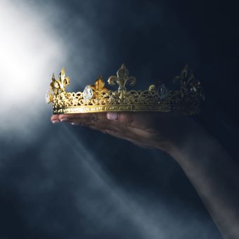 Hand holding an ornate gold crown with gemstones, against a dark, smoky background with a bright beam of light.
