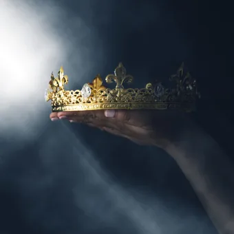 Hand holding an ornate gold crown with gemstones, against a dark, smoky background with a bright beam of light.