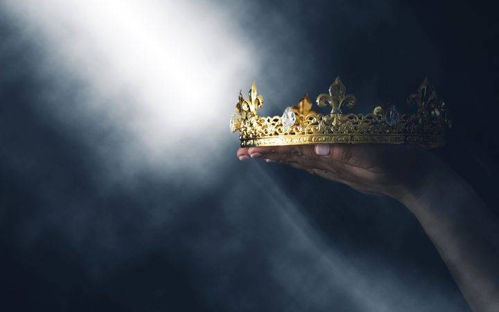 Hand holding an ornate gold crown with gemstones, against a dark, smoky background with a bright beam of light.