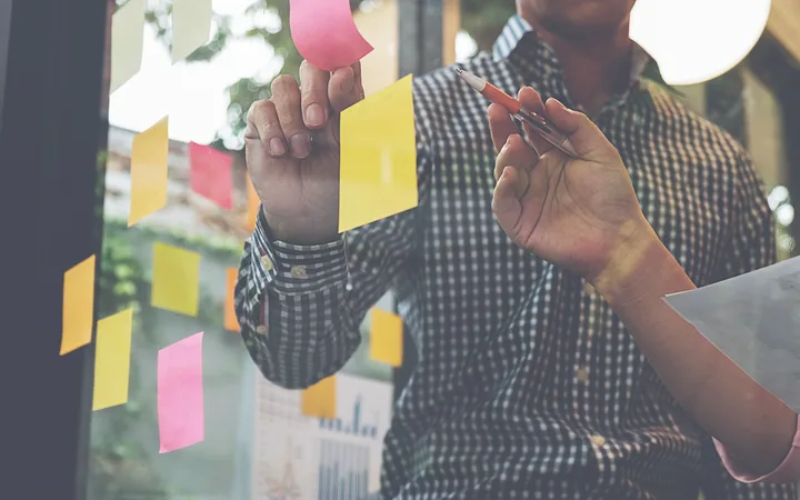 Person writing on colourful sticky notes on a glass wall, with charts and graphs visible in the background.
