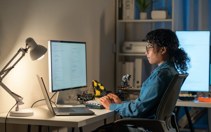 Person with curly hair and glasses sits at a desk coding, typing on a keyboard before a large monitor; a laptop, a lamp, and shelves with plants are visible.
