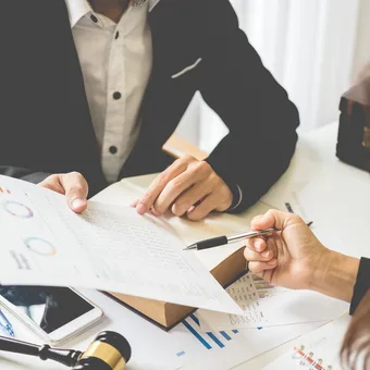 Two individuals exchanging documents and discussing financial charts in a business meeting at a desk with papers, a notebook, and a smartphone.