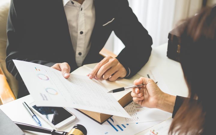 Two individuals exchanging documents and discussing financial charts in a business meeting at a desk with papers, a notebook, and a smartphone.