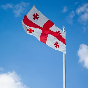A flag with a red cross and four smaller red crosses on a white background, waving against a blue sky with scattered clouds.