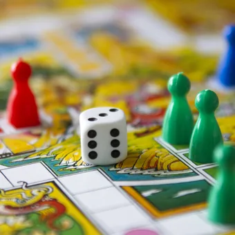 A close-up of a Monopoly game board with colorful plastic player pieces and a white die showing five.