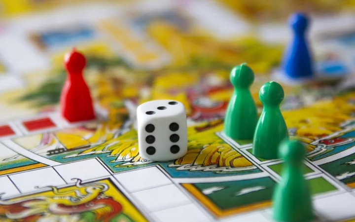 A close-up of a Monopoly game, showing a white die with five dots and coloured game pieces on a colourful game board.