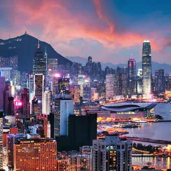 Hong Kong skyline at dusk with illuminated skyscrapers, Victoria Harbour, and mountains in the background. Bright city lights contrast with the evening sky.
