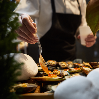 A person in a white shirt and black apron drizzles orange sauce over a platter of mussels with a glass spoon, on a wooden table.