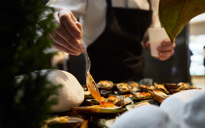 A person in a white shirt and black apron drizzles orange sauce over a platter of mussels with a glass spoon, on a wooden table.