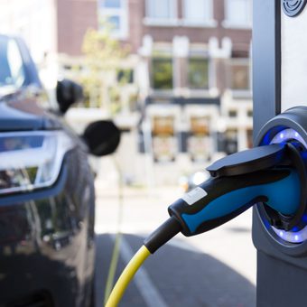 Close-up of an electric car charging: blue-and-black charging handle plugged into a port with a blue LED ring, city street blurred in the background.