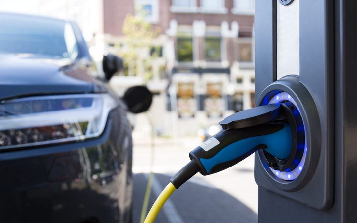 Close-up of an electric car charging: blue-and-black charging handle plugged into a port with a blue LED ring, city street blurred in the background.