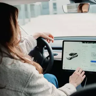 Woman driving a car while using a tablet mounted on the dashboard. The scene is inside a vehicle with buildings visible outside.