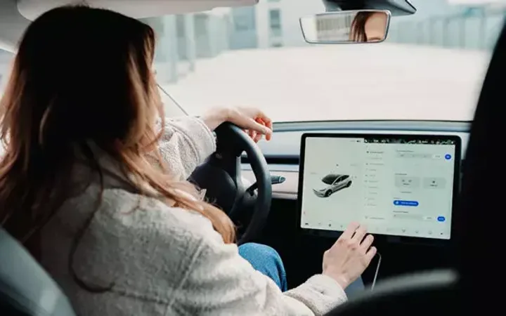 Woman driving a car while using a tablet mounted on the dashboard. The scene is inside a vehicle with buildings visible outside.