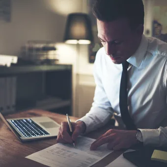 Man in white shirt and black tie reviewing documents at a desk with a laptop, in a dimly lit office with a city view through the window.