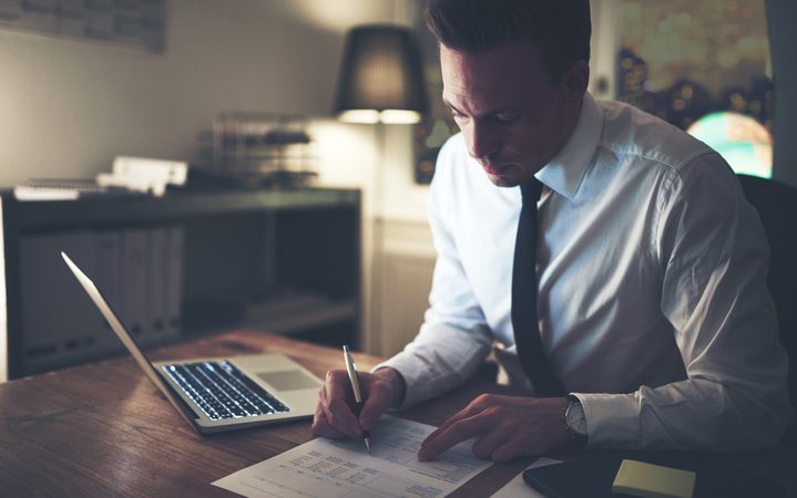 Man in white shirt and black tie reviewing documents at a desk with a laptop, in a dimly lit office with a city view through the window.