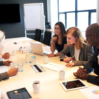 A diverse group of five professionals engaged in a meeting around a conference table, with laptops, tablets, and documents in a modern office setting.