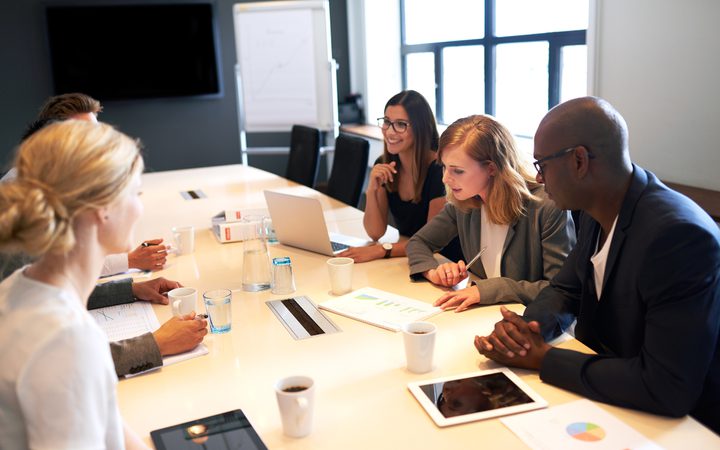 A diverse group of five professionals engaged in a meeting around a conference table, with laptops, tablets, and documents in a modern office setting.