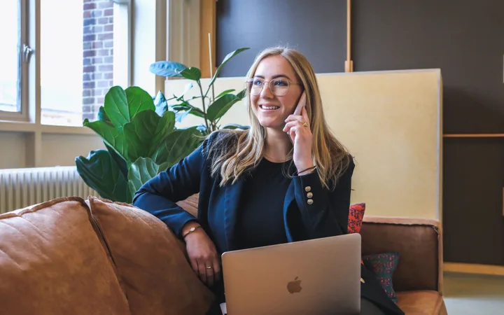 Person with long blonde hair, glasses, and a dark jacket sitting on a couch, talking on a mobile phone with a laptop on her lap in a bright, modern office.