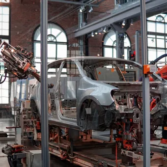 Robotic arms assemble a car frame inside a factory with large windows and exposed brick walls, showcasing automated manufacturing.