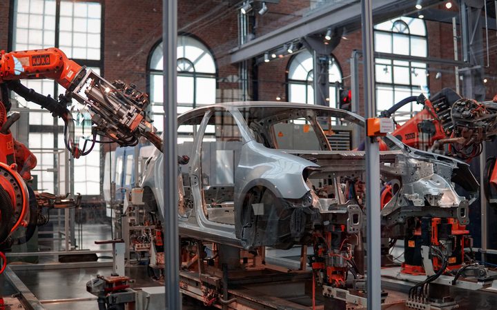 Robotic arms assemble a car frame inside a factory with large windows and exposed brick walls, showcasing automated manufacturing.