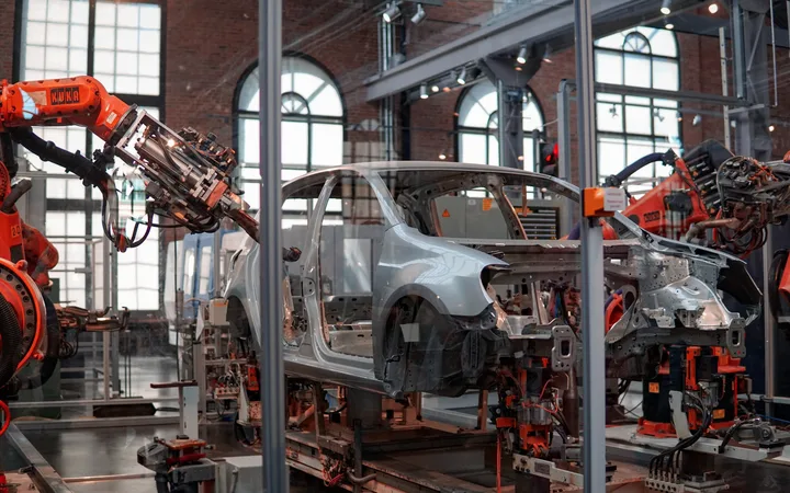Robotic arms assemble a car frame inside a factory with large arched windows and industrial equipment.
