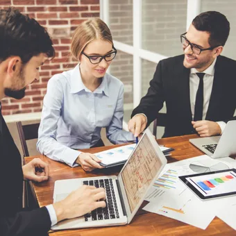 Four professionals in business attire collaborate at a desk with laptops, charts, and graphs, smiling and discussing data insights.