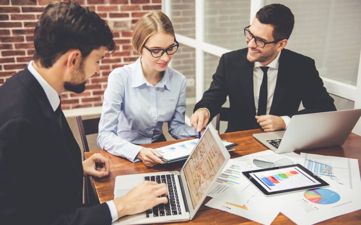 Four professionals in business attire collaborate at a desk with laptops, charts, and graphs, smiling and discussing data insights.