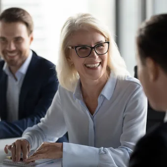 A woman with glasses, blonde hair, and a white shirt smiling and talking to a man in a suit during a business meeting, with a man in a suit smiling in the background.