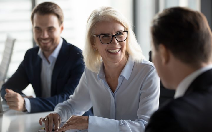 A group of three business professionals engaged in a friendly conversation, with a woman with blonde hair and glasses smiling warmly at a man, in a bright modern office.