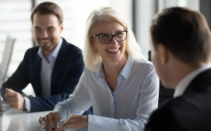 A group of three business professionals engaged in a friendly conversation, with a woman with blonde hair and glasses smiling warmly at a man, in a bright modern office.