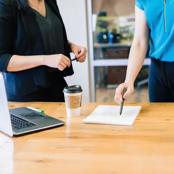 Two people are standing at a wooden table with a laptop, a notebook, and a takeaway coffee. One is holding a pen over the notebook, the other holding a marker.
