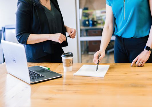 Two people are standing at a wooden table with a laptop, a notebook, and a takeaway coffee. One is holding a pen over the notebook, the other holding a marker.