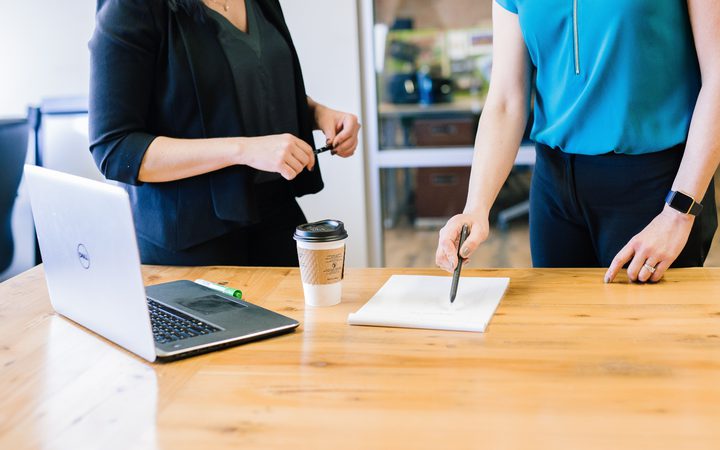 Two people are standing at a wooden table with a laptop, a notebook, and a takeaway coffee. One is holding a pen over the notebook, the other holding a marker.
