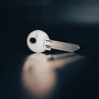 A single silver key lying on a dark surface, with light reflecting off it and its shadow cast on the surface.