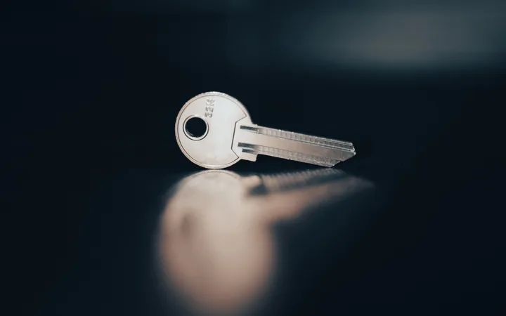 A silver key lies on a dark surface, with its reflection visible beneath it, illuminated softly against a dark background.