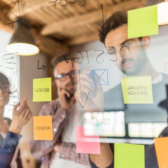 Portrait of a person in a blue suit, white shirt and polka-dot tie, wearing glasses with short hair and a trimmed beard, arms crossed against a grey gradient backdrop.