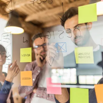 Portrait of a person in a blue suit, white shirt and polka-dot tie, wearing glasses with short hair and a trimmed beard, arms crossed against a grey gradient backdrop.