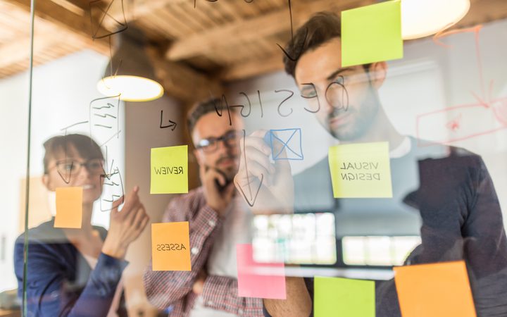 Three professionals collaborating and writing on a glass board with notes and sketches during a meeting in a modern office.