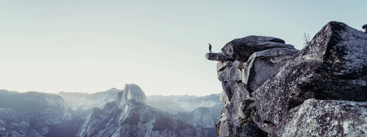 Personne debout sur un rocher au bord d'une falaise, avec des montagnes en arrière-plan et un ciel clair.