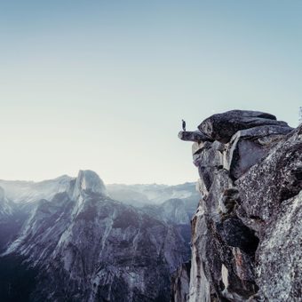 A person standing on a rocky ledge overlooking a mountain valley with peaks in the distance under a clear sky.