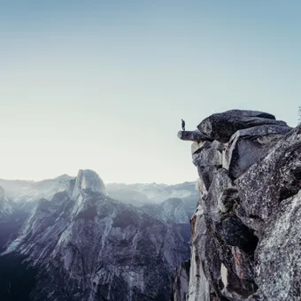 A person standing on a rocky ledge overlooking a mountain valley with peaks in the distance under a clear sky.