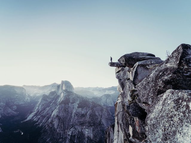 Personne debout sur un rocher au bord d'une falaise, avec des montagnes en arrière-plan et un ciel clair.