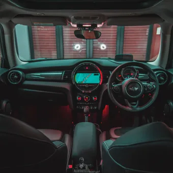 Inside a car with a central navigation screen, illuminated controls, and a steering wheel with a Mini logo, viewed from the back seats at night.