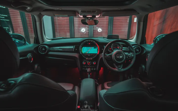 Inside a car with a central navigation screen, illuminated controls, and a steering wheel with a Mini logo, viewed from the back seats at night.
