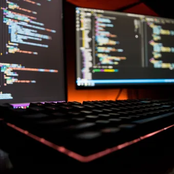 Dual monitors displaying colorful code in a dark room with a backlit keyboard in the foreground.