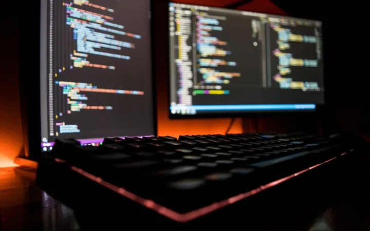 Two monitor screens displaying colourful lines of code with a backlit keyboard in the foreground, set against a dark, warm-toned background.