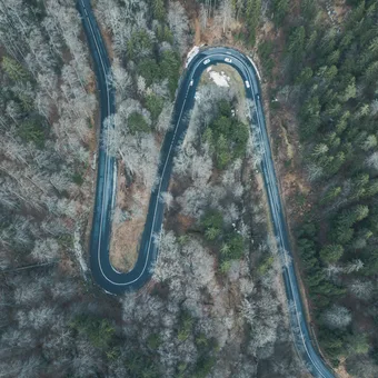 A winding mountain road through a snowy forest with bare and evergreen trees, viewed from above.
